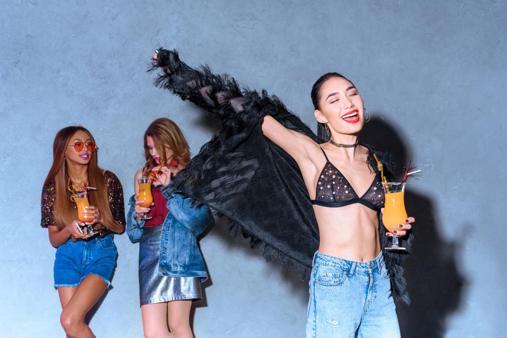 cheerful stylish asian girl holding glass with cocktail while friends drinking behind at party - Photo, Image