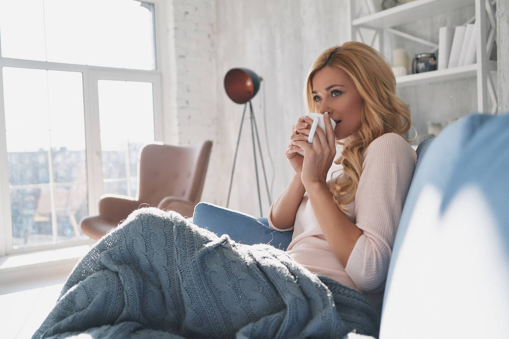Расслабленная и счастливая блондинка держит чашку и смотрит в сторону, сидя на диване дома
 - Фото, изображение