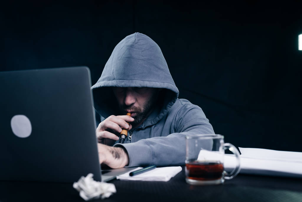 чоловік хакер в светрі з капюшоном сидить за ноутбуком і курить кальян
 - Фото, зображення