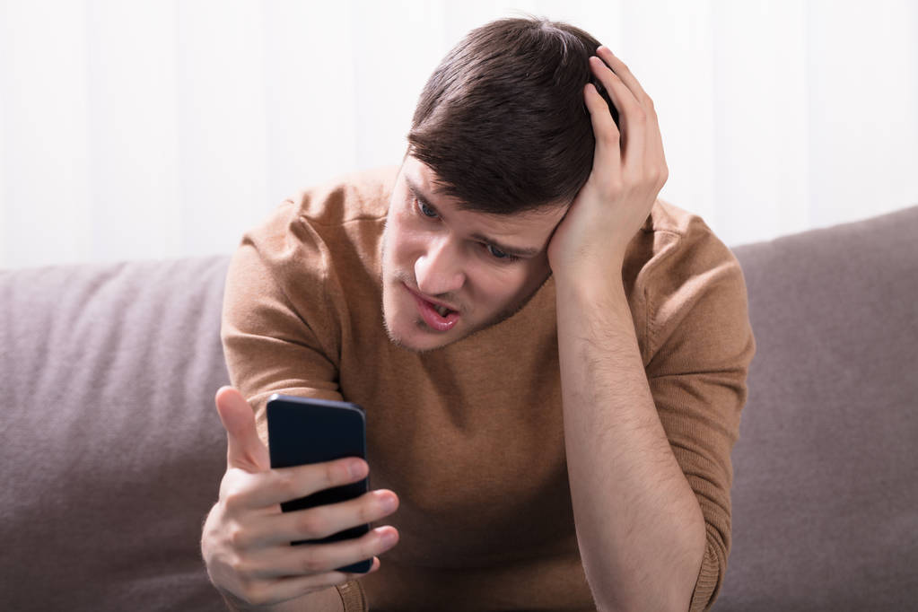 Shocked Young Man Sitting On Sofa And Looking At Smartphone At Home - Photo, Image