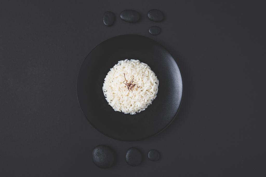 Blick von oben auf leckeren Reis auf Teller mit Kieselsteinen auf schwarzem Tisch - Foto, Bild