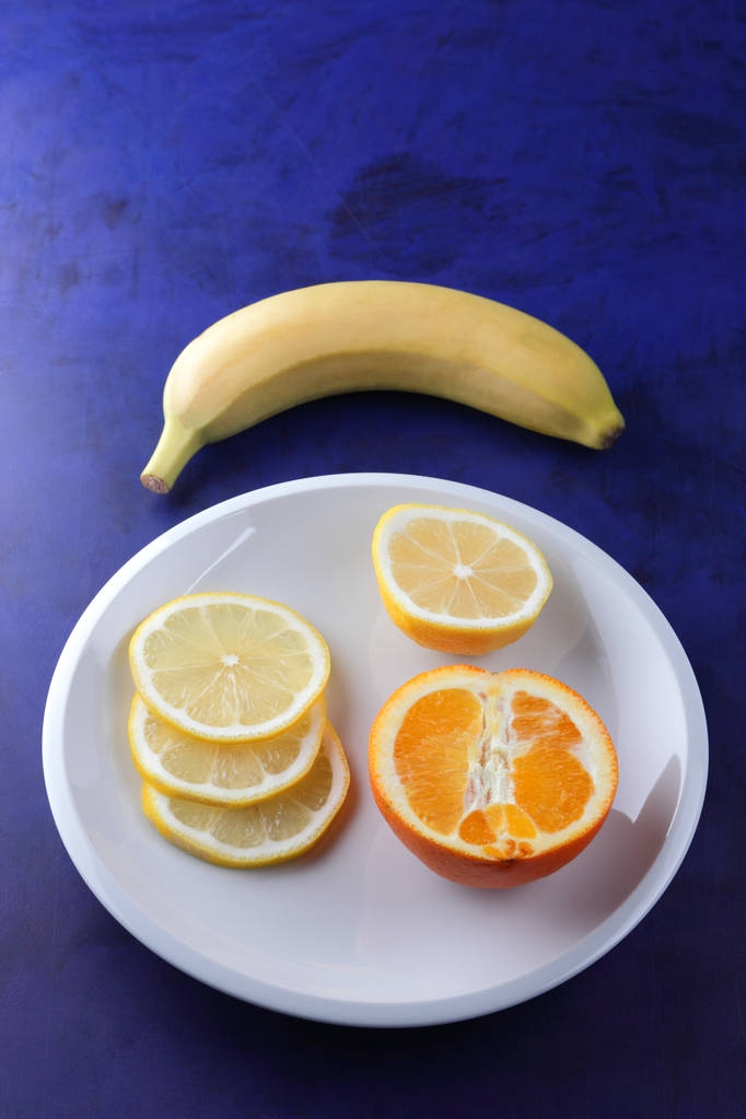 Цитрусовые на ультрафиолетовом фоне в стиле минимализма, ломтики лимона и полуоранжевого на белой тарелке, желтый банан на темно-синем фоне, здоровая еда, вегетарианская, поп-арт
 - Фото, изображение