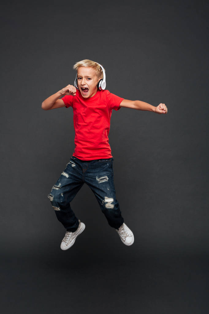 Image of excited little boy child jumping isolated over grey background. Looking camera listening music with headphones. - Photo, Image