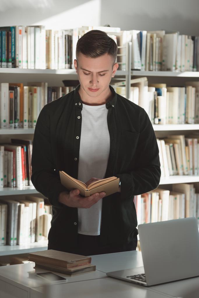 красивий студент стоїть і читає книгу в бібліотеці
 - Фото, зображення