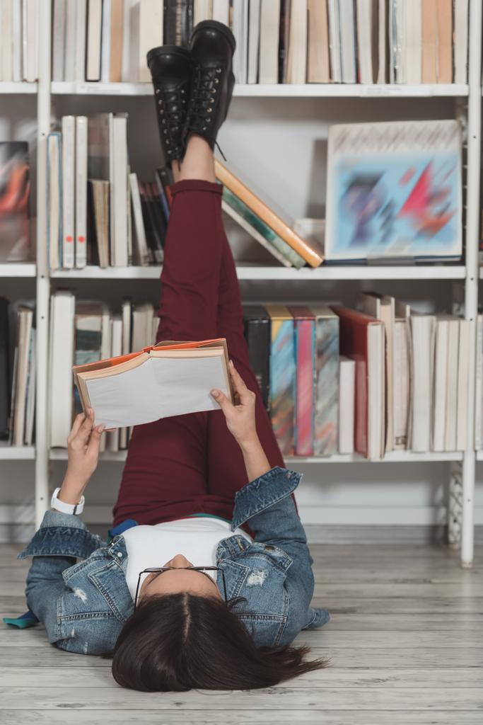Afrikanerin liegt mit erhobenen Beinen auf dem Boden in Bibliothek und liest Buch - Foto, Bild