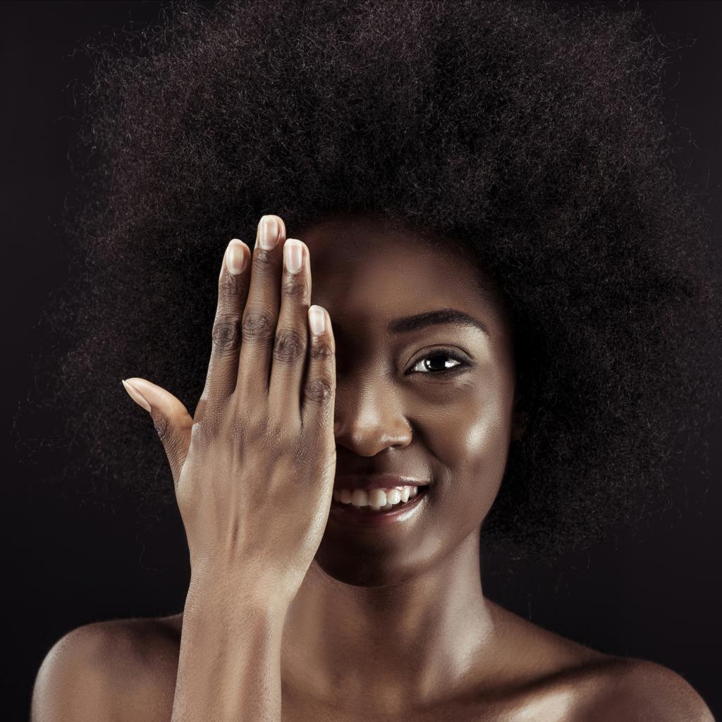 mujer afroamericana feliz que cubre el ojo con la mano aislada en negro
 - Foto, Imagen