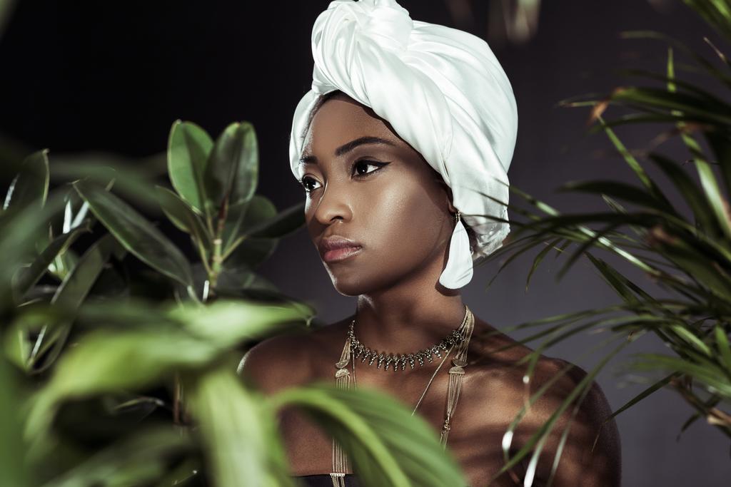 aantrekkelijke Afrikaanse Amerikaanse vrouw in de hoofd wrap stijlvolle draad achter bladeren - Foto, afbeelding