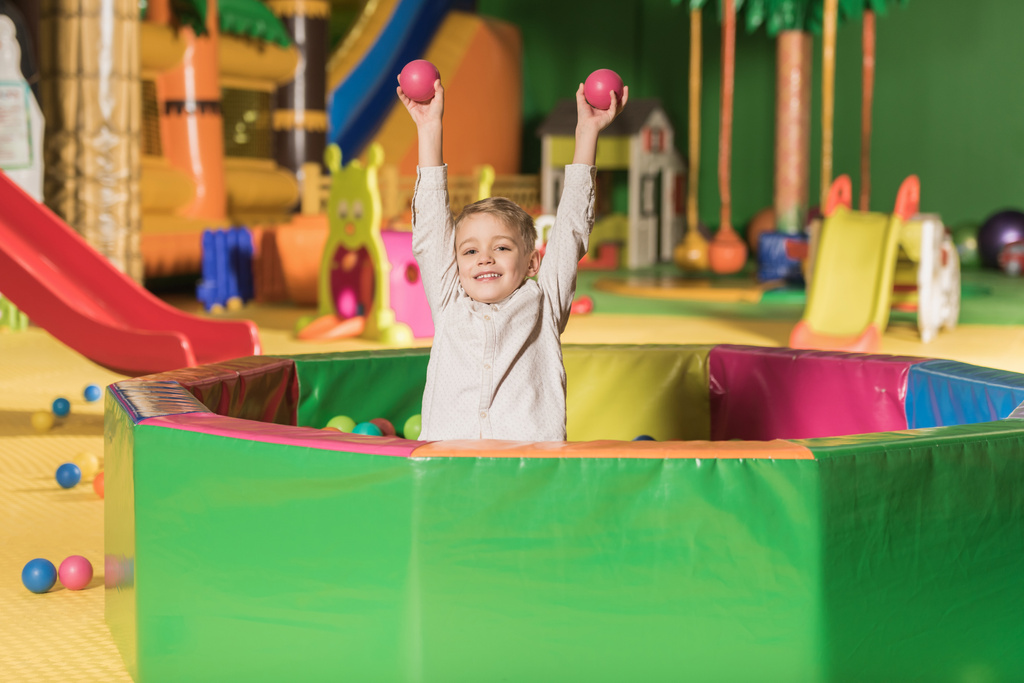αξιολάτρευτο μικρό αγόρι χαμογελά σε κάμερα ενώ παίζουν στην πισίνα με πολύχρωμες μπάλες  - Φωτογραφία, εικόνα