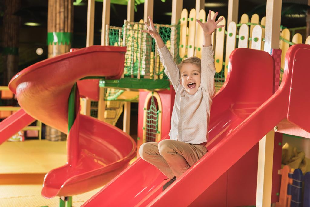 чарівний щасливий маленький хлопчик з піднятими руками грає на слайді в ігровому центрі
 - Фото, зображення