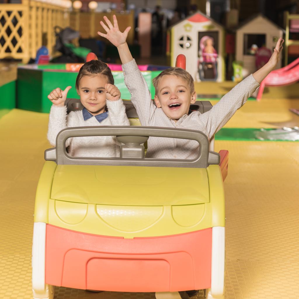 adorables frères et sœurs heureux assis dans la voiture jouet et souriant à la caméra dans le centre de divertissement
 - Photo, image