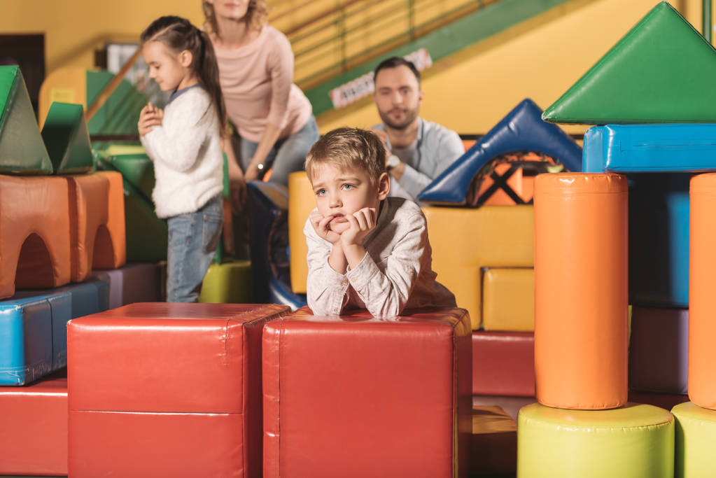 niño aburrido mirando hacia otro lado mientras la familia juega con bloques de colores en el centro de entretenimiento
 - Foto, Imagen