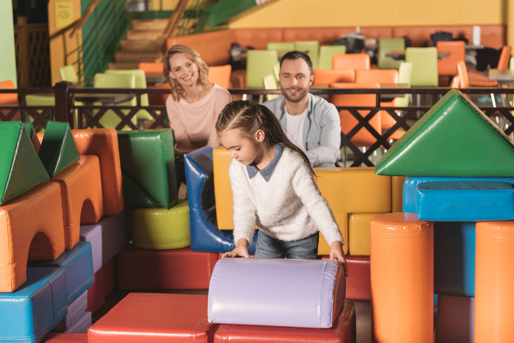 parents looking at cute little daughter building castle with colorful blocks in entertainment center - Photo, Image