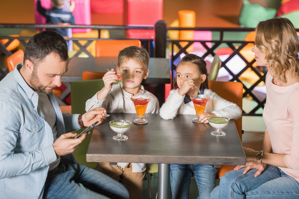 семья с двумя детьми едят вкусные десерты и отец с помощью смартфона в развлекательном центре
    - Фото, изображение