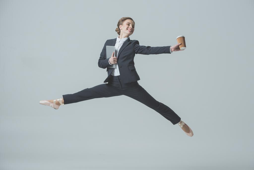 businesswoman in suit and ballet shoes jumping with coffee and tablet, isolated on grey - Photo, Image