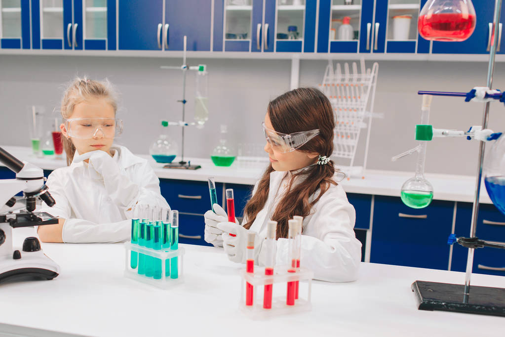 Двое детей в лабораторном халате учатся химии в школе. Молодые ученые в защитных очках проводят эксперименты в лаборатории или химическом шкафу. Изучение ингредиентов для экспериментов
 . - Фото, изображение