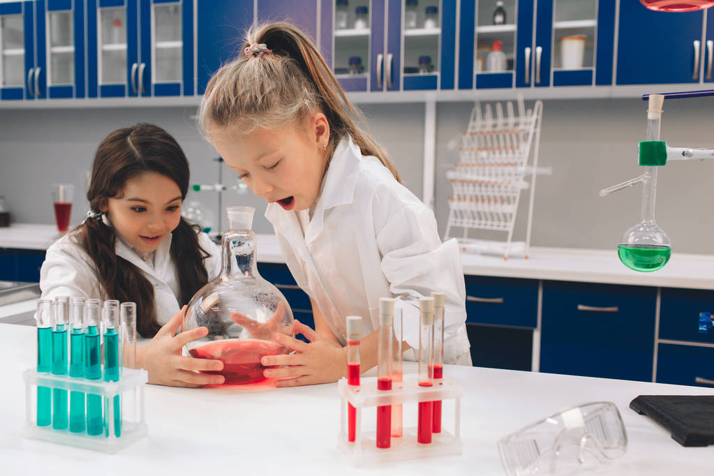 Dwa małe dzieci w fartuchu uczenia się chemii w szkole laboratorium. Młodych naukowców w ochronne okulary podejmowania eksperyment w laboratorium lub gabinetu chemicznego. Badanie składników do eksperymentów . - Zdjęcie, obraz