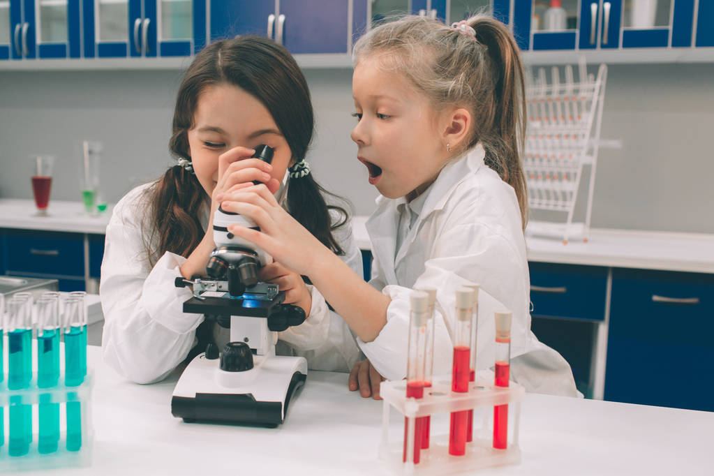 Zwei kleine Kinder im Laborkittel lernen Chemie im Schullabor. Junge Wissenschaftler in Schutzbrillen experimentieren im Labor oder Chemiekabinett. Blick durchs Mikroskop - Foto, Bild