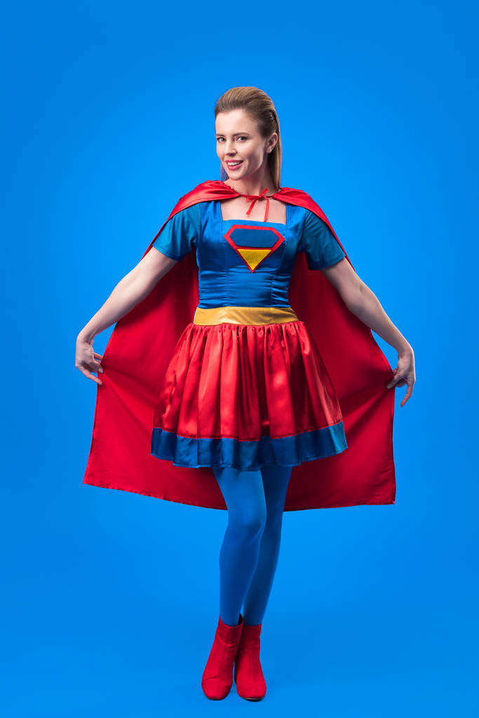 atrakcyjna kobieta w strój superbohatera, trzymając cape na niebieskim tle - Zdjęcie, obraz