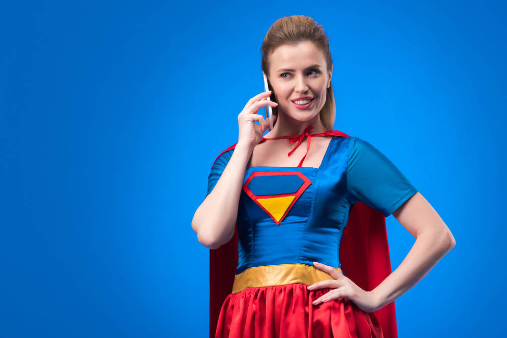portrait of smiling woman in superhero costume talking on smartphone isolated on blue - Photo, Image