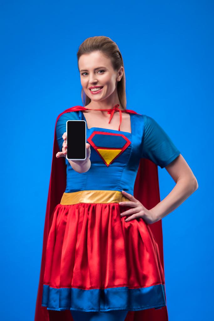 πορτρέτο του χαμογελαστό γυναίκα στο κοστούμι superhero δείχνει smartphone με κενή οθόνη που απομονώνονται σε μπλε - Φωτογραφία, εικόνα