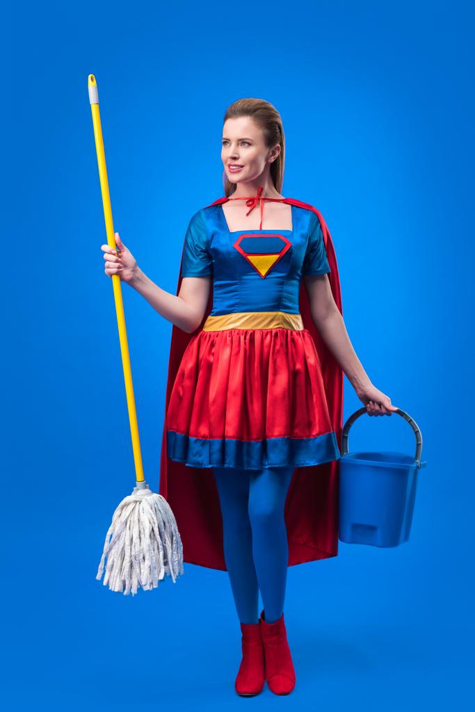 attraktive Frau im Superheldenkostüm mit Wischmopp und Eimer zum Putzen isoliert auf blau - Foto, Bild