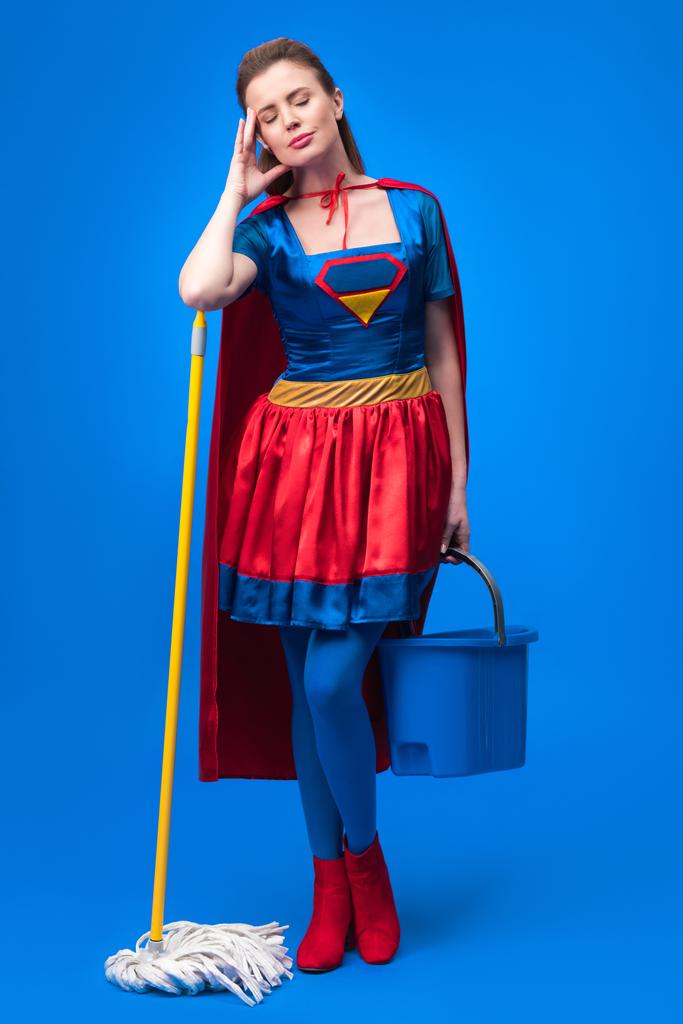 unavená žena v kostýmu superhrdiny s mop a kbelík pro čištění, izolované na modré - Fotografie, Obrázek
