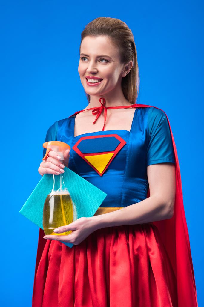 Portret van lachende vrouw in superheld kostuum met doek en schoonmaakmiddel in handen geïsoleerd op blauw - Foto, afbeelding