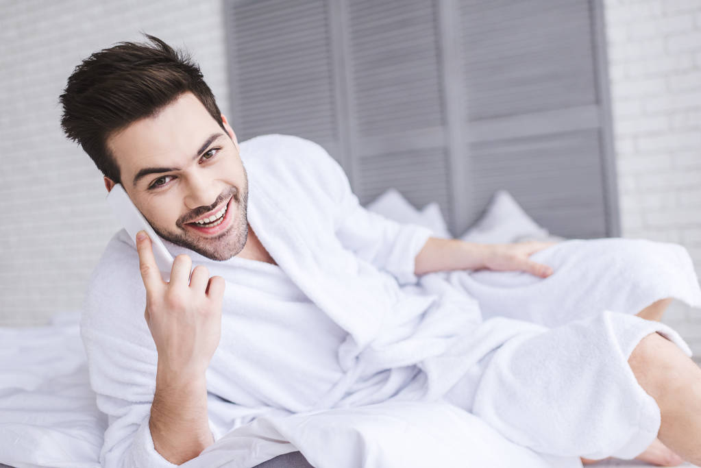 handsome young man in bathrobe talking by smartphone and smiling at camera  - Photo, Image