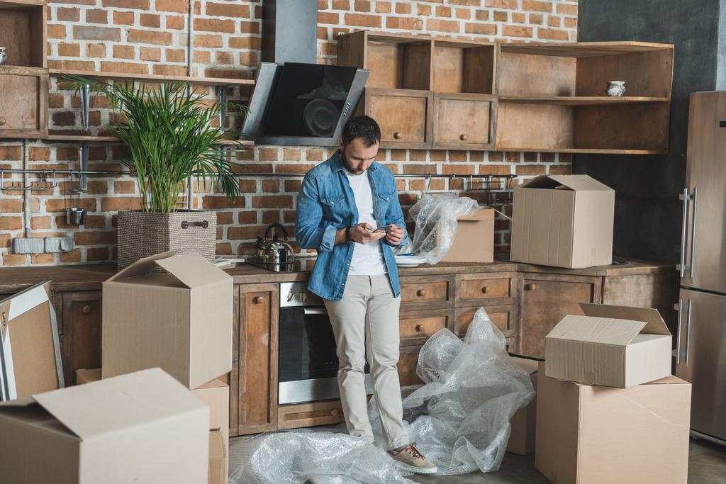 hombre usando smartphone mientras está de pie entre cajas de cartón en nuevo apartamento
 - Foto, imagen
