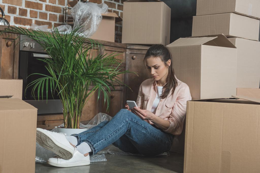 jovem mulher usando smartphone enquanto se senta entre caixas de papelão em novo apartamento
 - Foto, Imagem
