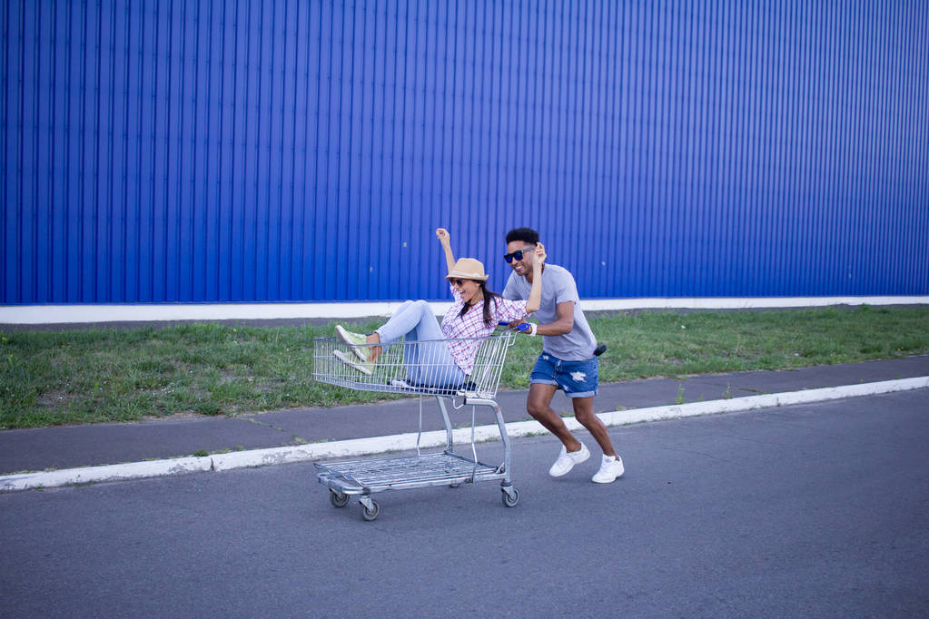 Young hipsters couple riding on trolley on empty shop parking during sunrise and have good funny time - Photo, Image