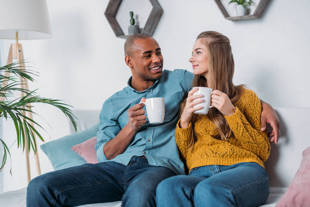 Multikulturelles Paar sitzt mit Kaffee auf Sofa und schaut sich an - Foto, Bild
