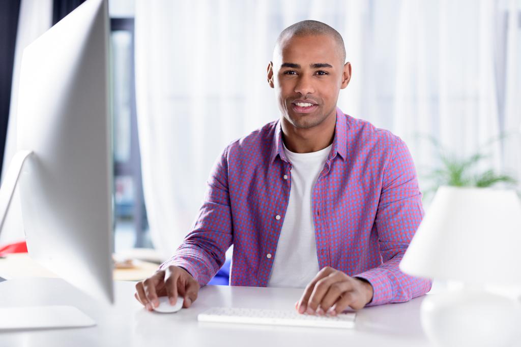 sonriente afroamericano hombre sentado en el ordenador y mirando a la cámara
 - Foto, imagen
