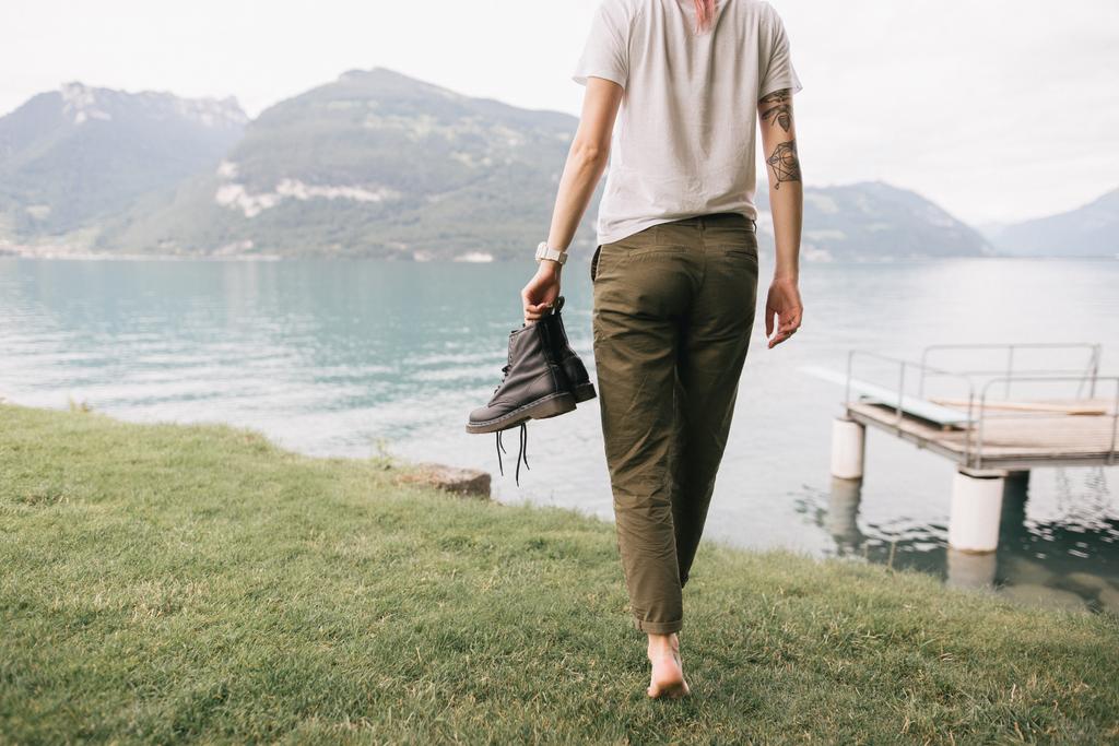 обрізаний знімок дівчини, що тримає взуття і ходить босоніж на траві біля красивого гірського озера
 - Фото, зображення
