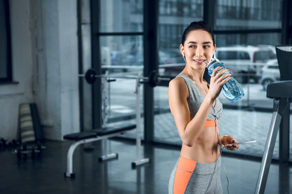 νεαρή γυναίκα σπορ πόσιμο νερό και κοιτάζοντας πίσω στο γυμναστήριο - Φωτογραφία, εικόνα