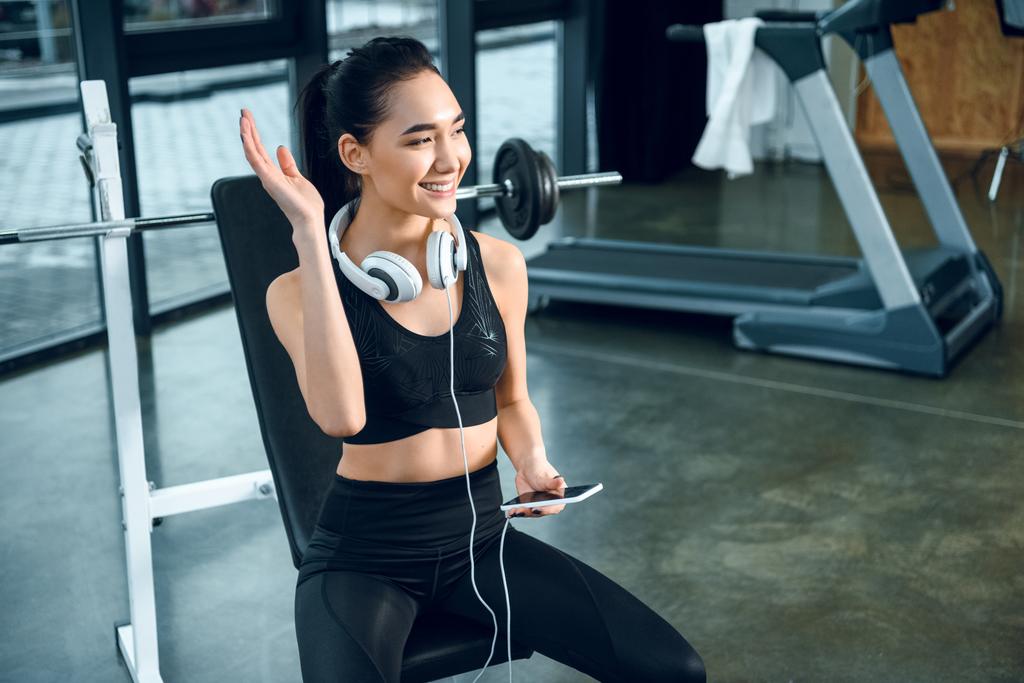 junge, fitte Frau entspannt sich im Fitnessstudio mit Smartphone und winkt jemandem zu - Foto, Bild