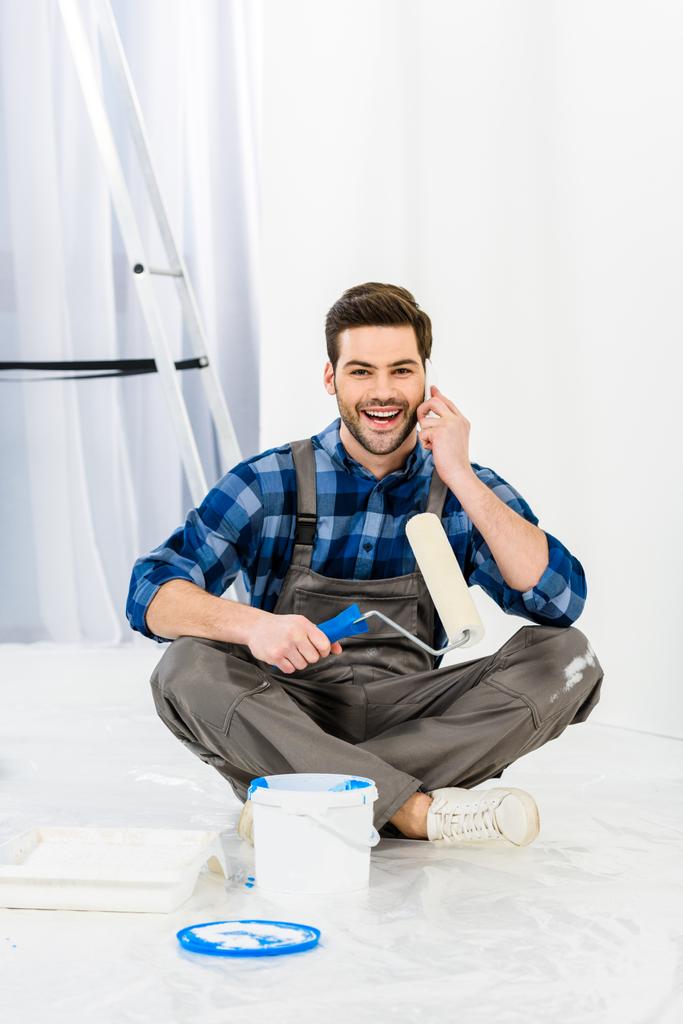hombre sonriente sentado con pincel de rodillo de pintura y hablando por teléfono inteligente
 - Foto, imagen