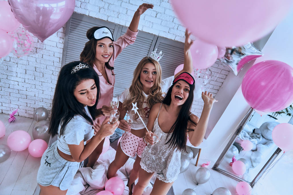 Вид сверху на четырех привлекательных улыбающихся молодых женщин, устраивающих вечеринку в спальне с алкоголем и воздушными шариками
 - Фото, изображение