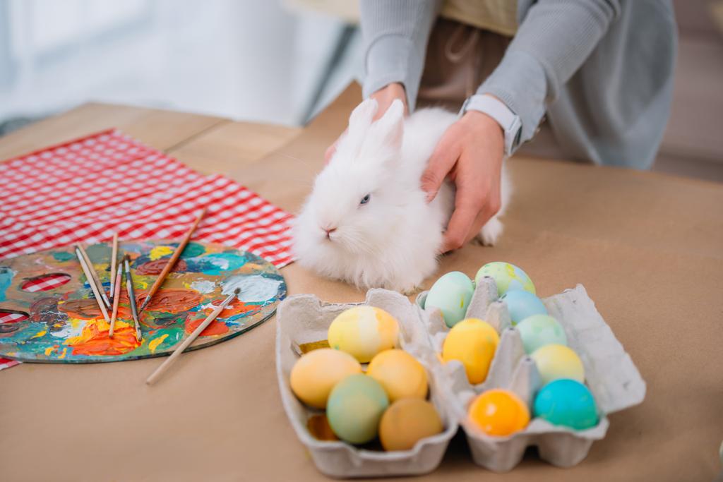 женщина с белым кроликом на столе с разноцветными пасхальными яйцами
 - Фото, изображение