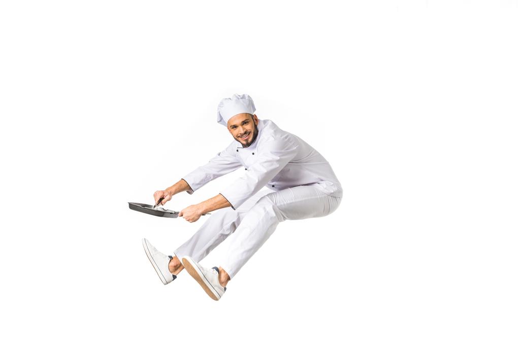 Gelukkig jonge chef-kok met koekenpan springen en kijken naar camera geïsoleerd op wit   - Foto, afbeelding