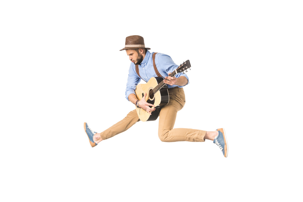 pohledný mladý hudebník v klobouku hrát na kytaru zatímco skákání izolované na bílém - Fotografie, Obrázek