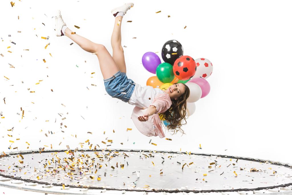 smiling girl holding colorful balloons and falling on trampoline isolated on white  - Photo, Image