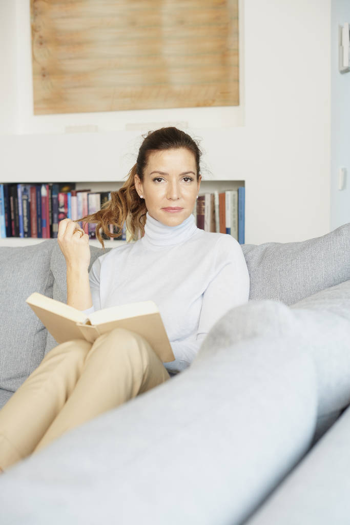 Szép mosolygó érett nő ül a kanapén és olvasókönyv otthon.  - Fotó, kép