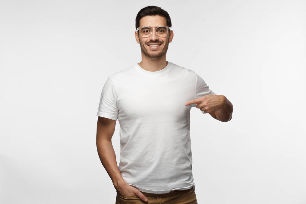 Привабливий молодий чоловік в прозорих окулярах, вказуючи на чисту білу сорочку з індексним пальцем, показує порожній простір для вашого рекламного тексту або логотипу, заявляючи, що ізольований на сірому фоні
 - Фото, зображення