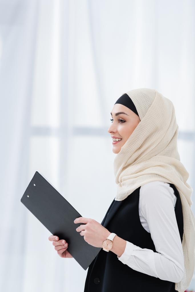 Seitenansicht einer lächelnden arabischen Geschäftsfrau mit Ordner in der Hand im Büro - Foto, Bild