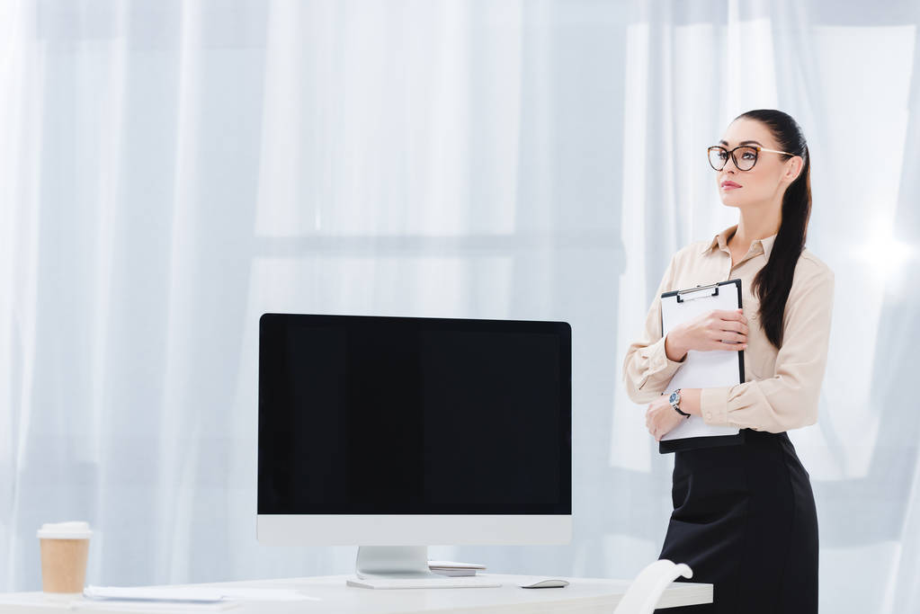 atractiva mujer de negocios con bloc de notas de pie en el lugar de trabajo con pantalla de ordenador en blanco
 - Foto, Imagen