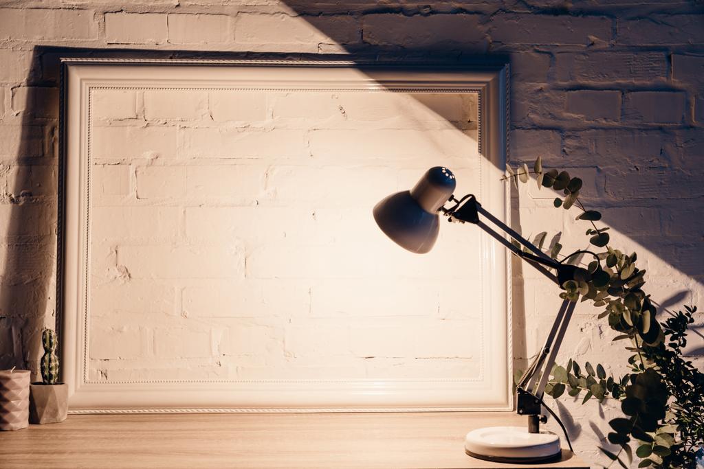 table lamp illuminating white empty brick wall with frame, mockup concept - Photo, Image