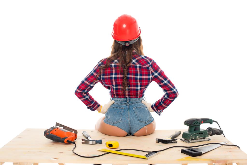 vista trasera de chica sexy en hardhat sentado en mesa de madera con herramientas, aislado en blanco
 - Foto, Imagen
