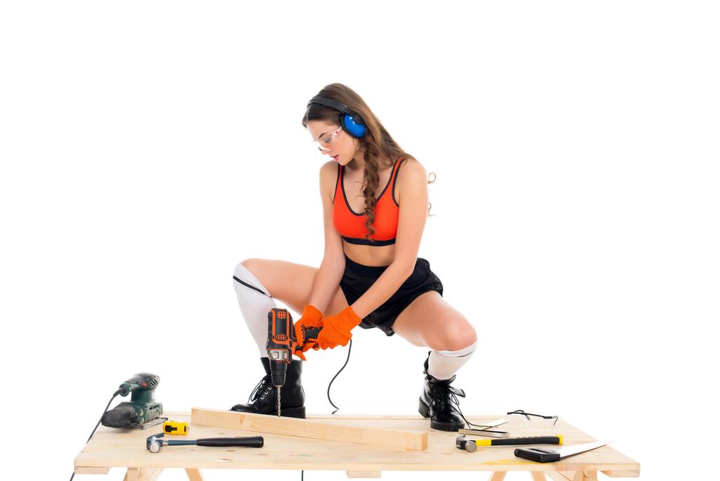 piękna dziewczyna w słuchawki ochronne, praca z wiertarka elektryczna w drewniany stół z narzędziami, na białym tle - Zdjęcie, obraz
