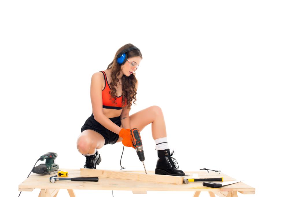 chica sexy en auriculares protectores que trabajan con taladro eléctrico en la mesa de madera con herramientas, aislado en blanco
 - Foto, imagen